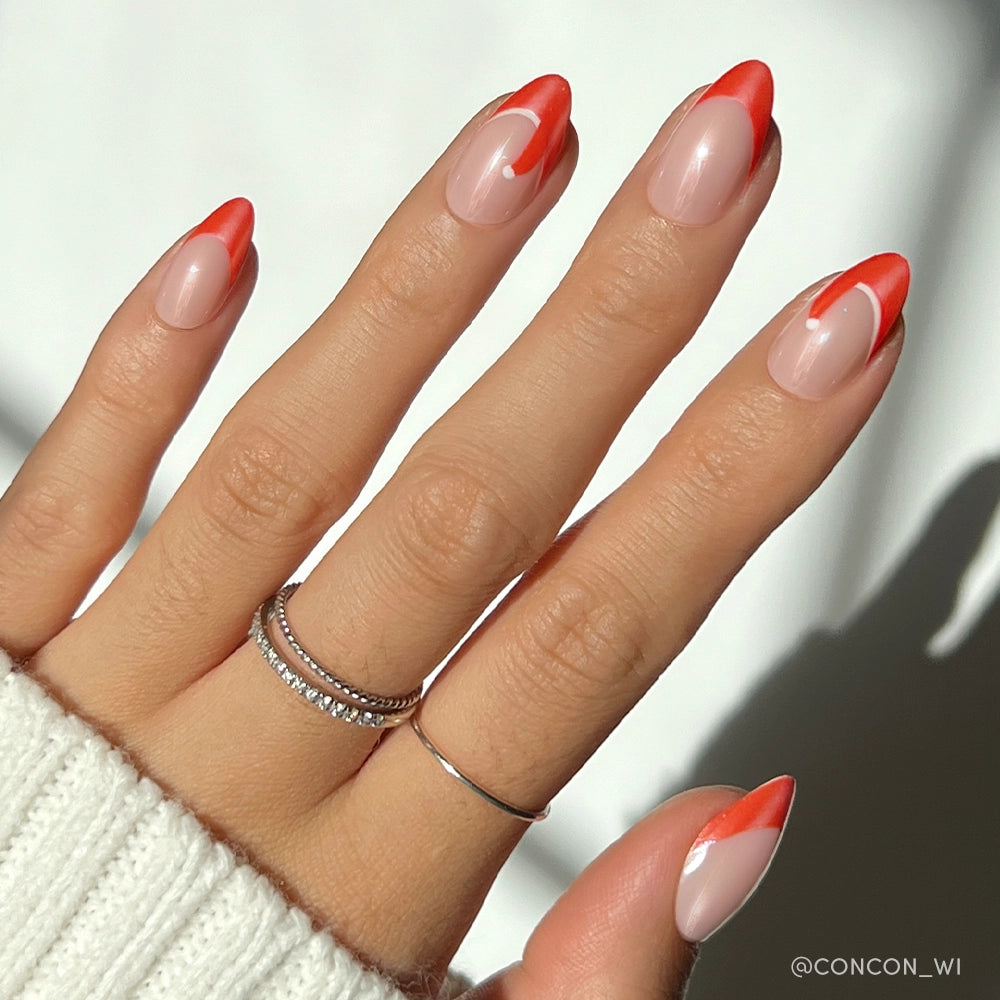 A hand with glamnetic’s Mrs. Claus glazed almond nails—nude base and red-orange abstract tips—adorned with two silver rings and a white knit sweater sleeve, creating a chic, festive look.